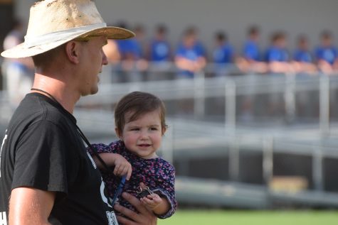 ITS FOOTBALL BABY: Holding his daughter Camilla, head football coach Thomas Gammerdinger runs the football teams at the Manor scrimmage in 2019. At this particular game, Gammerdinger had to bring his daughter in spite of the summer heat because she hadnt hadnt been feeling well. Luckily, he had plenty of help from his assistant coaches. Camillas personal favorite being Coach Steve Searle. Anytime Camilla is up here, theyll all jump in and help take care of her, Gammerdinger said.Though Camilla was a fun addition to the team on Thursday, she is seldom on the sidelines at games. Gammerdinger says his schedule with football limits his time with his daughter: Its hard not being with her all the time, he said. I usually wake up and leave before she even wakes up, then I come home to her fast asleep. Although it is never a good thing for a baby to be sick, parents and players at McCallum were sure glad that they got to see her at the scrimmage. 
