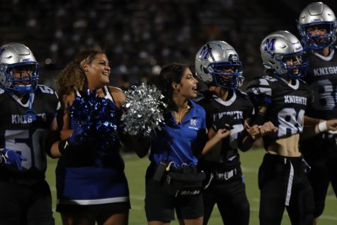 Seniors Genesis Ritchardson and Ximena de la Garza link pinkies during McCallum homecoming game. Both are members of the color guard while also balancing a second football season extracurricular: Ritchardson is a member of the cheer team and de la Garza is an athletic trainer. 