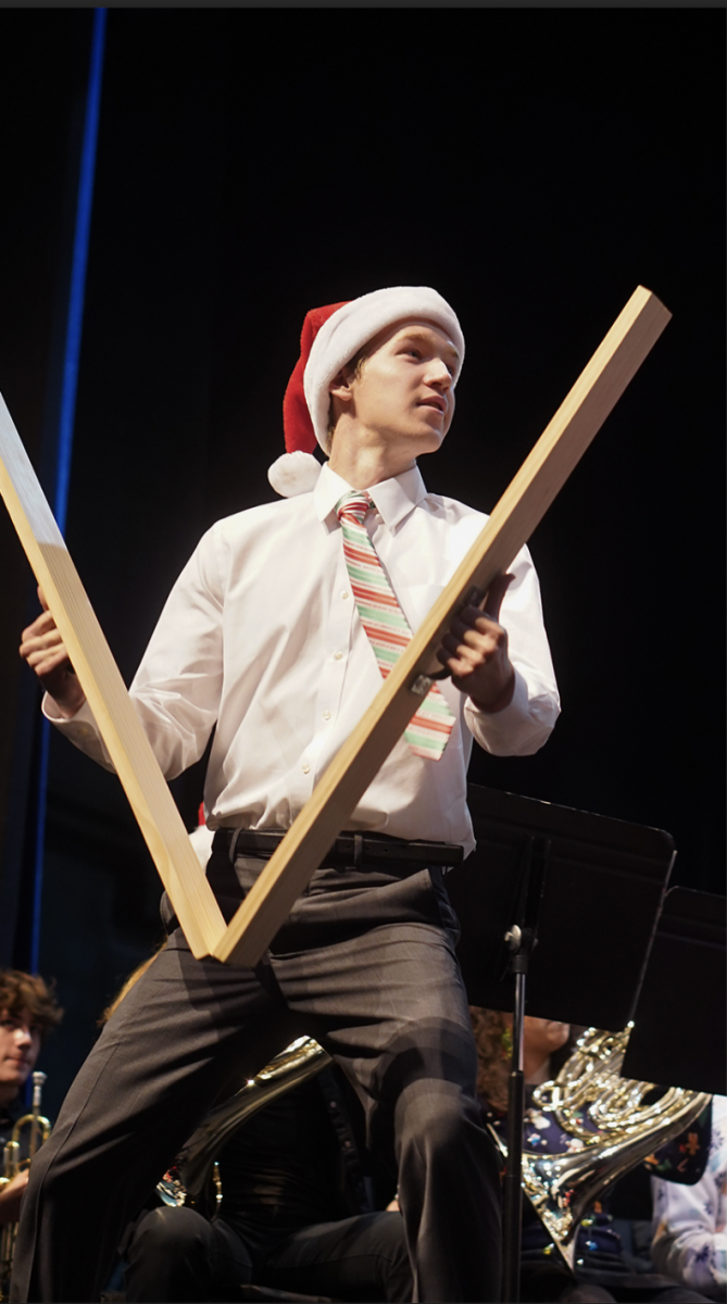 A KNEE-SLAPPING BIT: Senior percussionist Thomas Ross performs with the slapstick in ‘Sleigh Ride’ at the band’s winter concert last Tuesday. The slapstick is a less common percussion instrument often used in holiday music events. 

“The percussion section used multiple different sized slapsticks as a funny bit for the audience, coming out of nowhere with a bigger slapstick than the last,” Ross said. “Though I only get about one or two notes throughout the entire piece, it’s still a fun instrument to play.”

The concert consisted of holiday songs and a holiday-themed percussion ensemble. 

“The main focus is just playing together and having fun,” Ross

The use of the slapstick and “simplicity” of the winter show are examples of the concert season being, for band members like Ross, less intense than the marching season. 

“Concert season is the fire after a long winter,” Ross said.

While preparing for concerts still requires hours in rehearsal dedicated to a multitude of ensemble and solo pieces, Ross still feels that it is not as stressful as being in the midst of football halftime performances and day-long marching competitions. 

“Concert season is not as demanding and is laid back for the most part,” Ross said. 

Caption by Francie Wilhelm. 