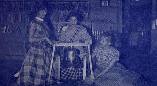 Laneta Stefka (Knight Editor), Ann Kinch (Blue Brigade President), and Jo Eickmann (Shield Editor) admire the Victory Bell in its habitat of last year, the Travis High library. Eickamann is pointing out a previous McCallum victory which the Knights hope to duplicate in tonights McCallum-Travis game. Caption and photo by 1957 Shield staff. 